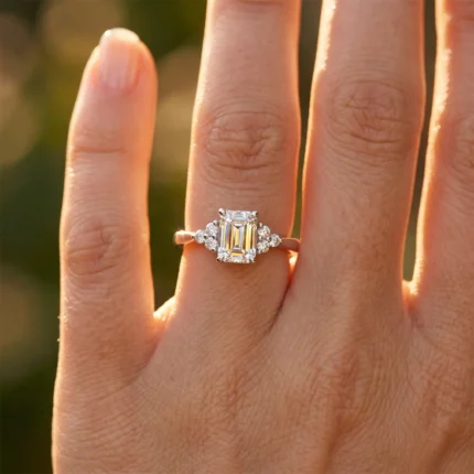 Close-up of a finger wearing an emerald-cut diamond ring with small side stones on a gold band, illuminated by warm sunlight.