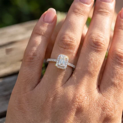 Close-up of a woman's left hand wearing a silver diamond ring with a rectangular emerald-cut center stone surrounded by a halo of smaller diamonds, in sunlight.